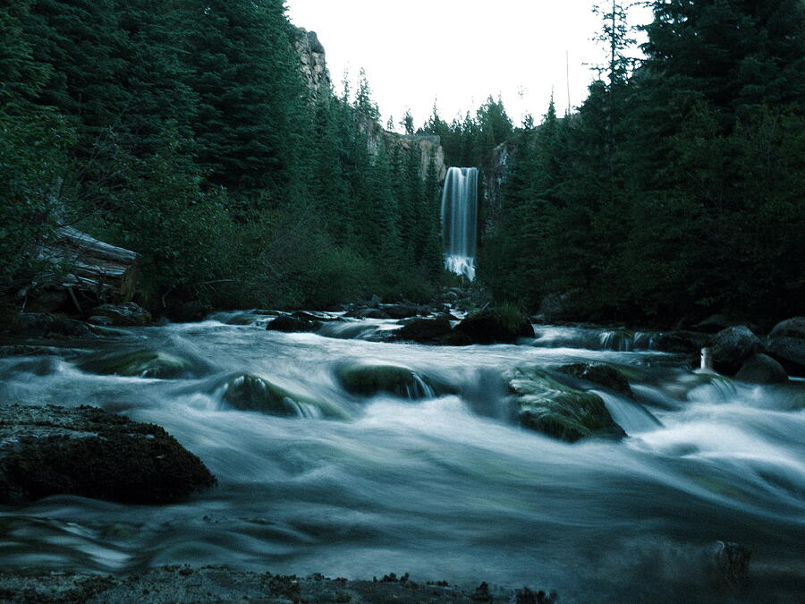 A waterfall framed by dark forest, with flowing water over rocks in the foreground.