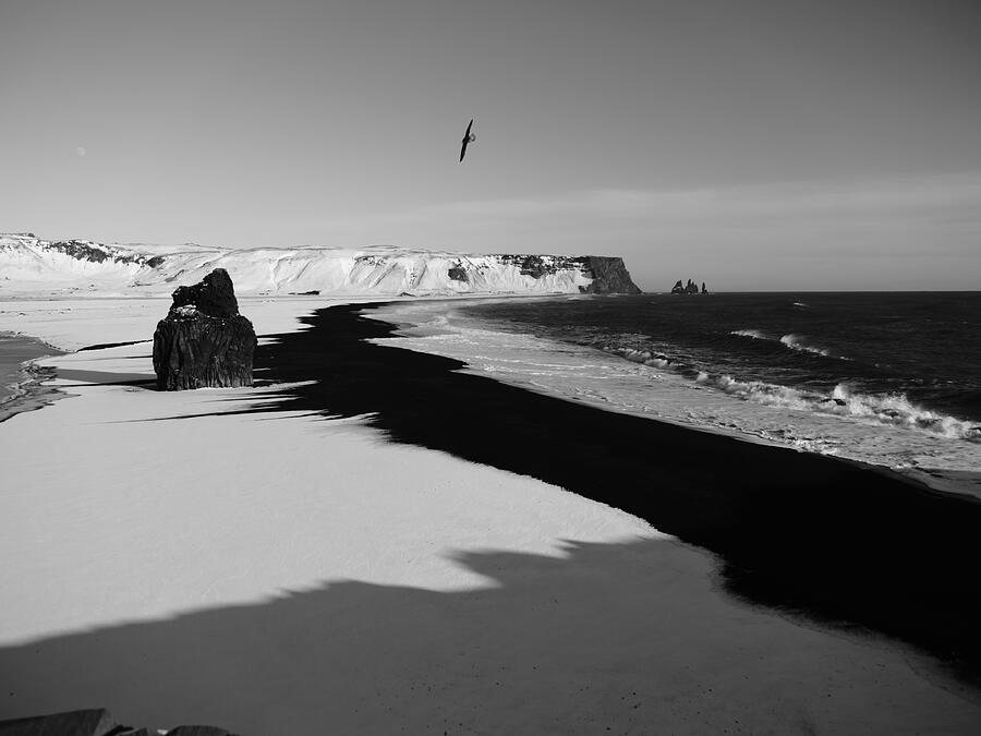 Snow and black sand meet dark ocean water, photographed in black and white.