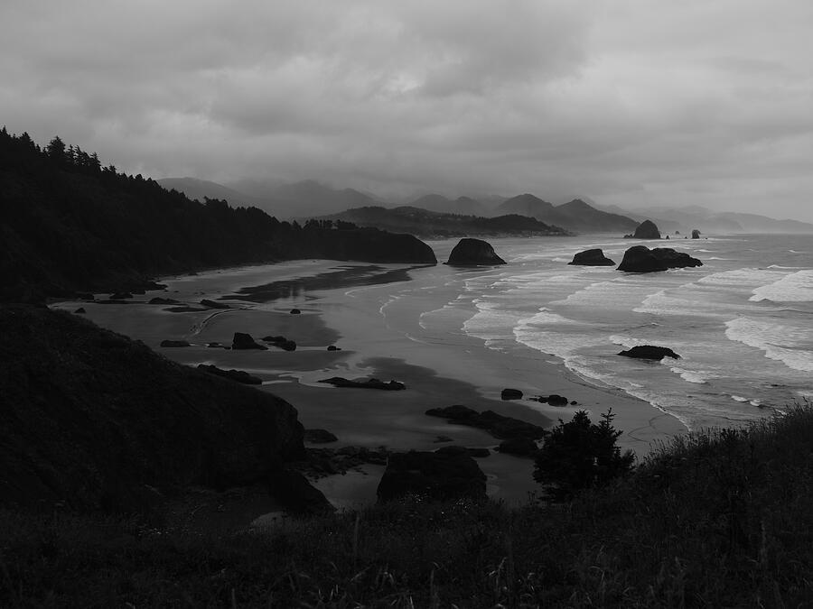Sea stacks, pale surf, and dark coastal forest beneath an overcast sky, in black and white.