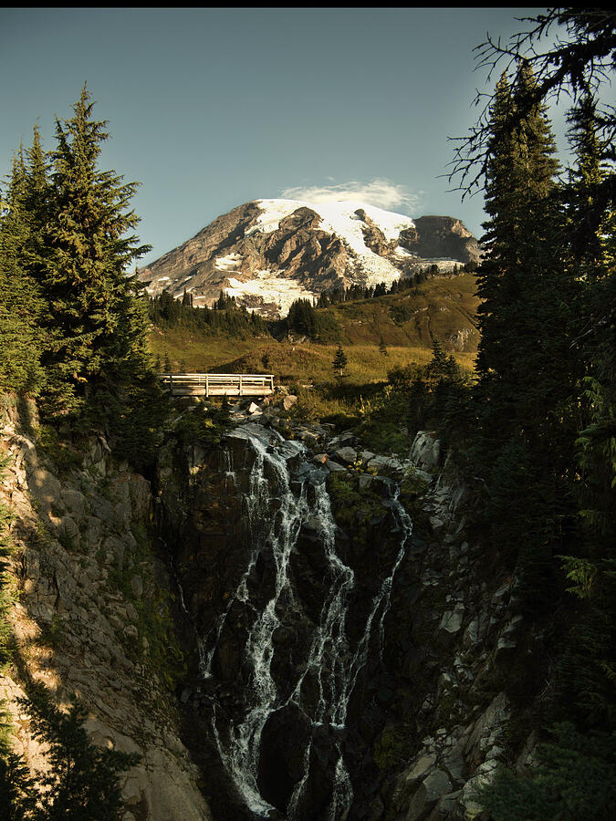 A waterfall drops through a wooded gorge beneath a snow-capped mountain.